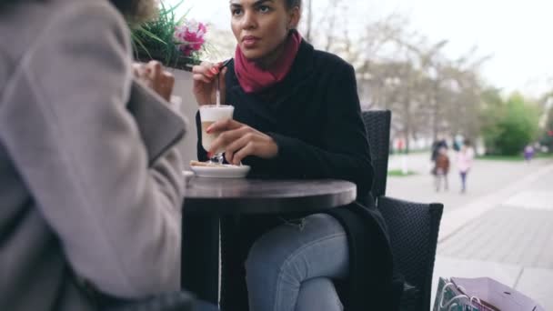 Tilt up of Two attractive mixed race women talking and drinking coffee in street cafe. Friends have fun after visiting mall sale - Metraje, vídeo