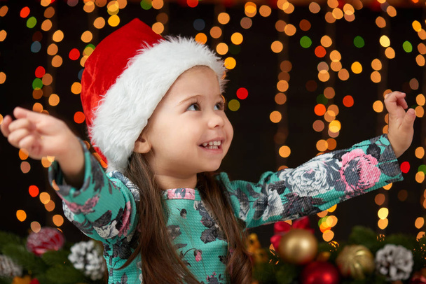 Kindermädchenporträt in Weihnachtsdekoration, fröhliche Emotionen, Winterferienkonzept, dunkler Hintergrund mit Beleuchtung und Bokelichtern - Foto, Bild