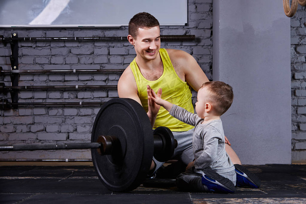 Young sporty father and little cute son sitting near barbell against brick wall in the cross fit gym. - 写真・画像