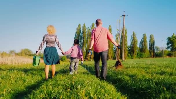 Back view: A family of farmers with a small son go together to plant a tree. Bear the apple tree seedlings, shovel and watering pad. Steadicam shot, side view - Záběry, video