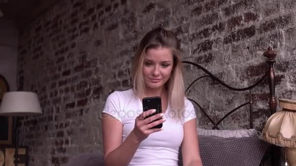 Blonde women using smart phone sitting on the bed. - Materiał filmowy, wideo