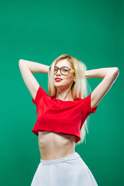 Блондинка дівчина з довгим волоссям і окулярів носіння біла спідниця і короткий Топ червоний є постановки в студії на зелений фон. - Фото, зображення