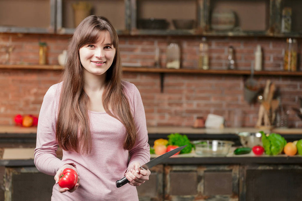 A woman in the kitchen near the table holds a tomato and a knife, and on the table lie fresh vegetables - Foto, Imagen