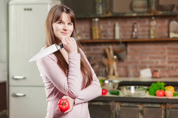 Жінка на кухні біля столу тримає помідор і ніж, а на столі лежать свіжі овочі
 - Фото, зображення