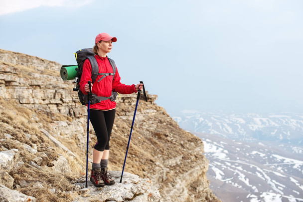 A slender girl in a cap with sticks for Nordic walking with a backpack and a folded rug for relaxation stands in the mountains against the backdrop of rocks and distant lands - Фото, изображение