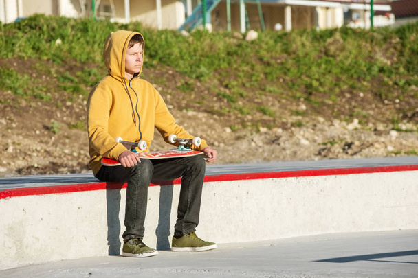 Young guy sitting in a skate park looking out into the distance and holding a skateboard - Photo, image