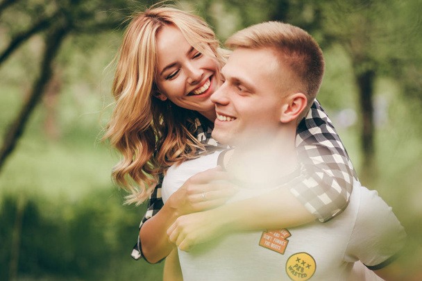 Ήπια οικογενειακή φωτογραφία της όμορφο νεαρό ζευγάρι. Στέκονται σε φόντο της apple στον κήπο. Η γυναίκα αγκαλιάζει ο άντρας από πίσω και τον κοιτάζει και χαμογελάει. Εκείνη στην πουκάμισο καρό, το λευκό T-shirt - Φωτογραφία, εικόνα