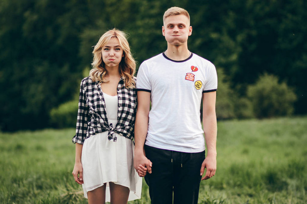 Young beautiful couple stands on a green park background. They hold hands. The blonde is dressed in a light white dress, and on the guy is a white T-shirt. The couple is crooked and fooling around. - Foto, imagen