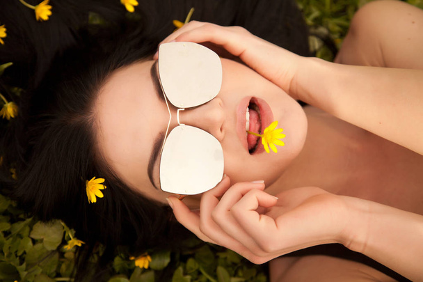Yaz aylarında sarı çiçeklerle çim üzerinde yalan gümüş ayna gözlüklü genç ve güzel esmer - Fotoğraf, Görsel