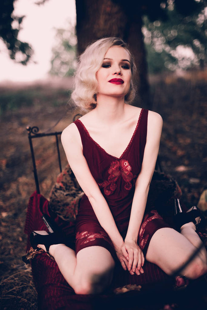 Kaunis ja tyylikäs blondi nainen punaiset huulet ja hiukset aallot yllään viini punainen yöpaita poseeraa sängyssä ulkona syksyllä, retro vintage tyyli ja muoti
. - Valokuva, kuva