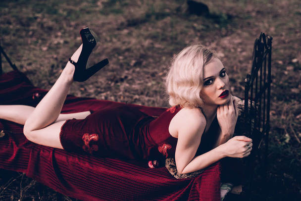 Красива і елегантна блондинка з червоними губами і хвилями волосся у вині червона сукня позує на ліжко на відкритому повітрі осінь, ретро вінтажний стиль і мода
. - Фото, зображення