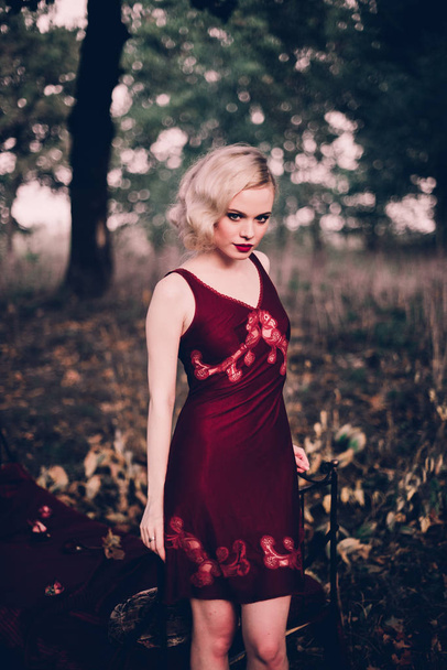 Beautiful and elegant blonde woman with red lips and hair waves wearing wine red nightie posing on the bed outdoors autumn, retro vintage style and fashion. - Foto, afbeelding