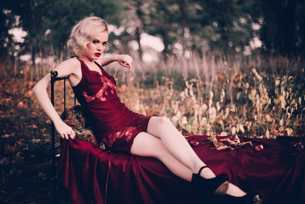 Mulher loira bonita e elegante com lábios vermelhos e ondas de cabelo vestindo vinho camisa vermelha posando na cama ao ar livre outono, estilo vintage retro e moda
. - Foto, Imagem