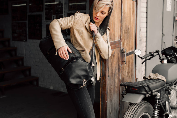 Een jonge vrouw met een leerzak in de buurt van de garage. Een vrouw in een lederen jas. Motorfiets in de buurt van de garage. Lederen tas - Foto, afbeelding