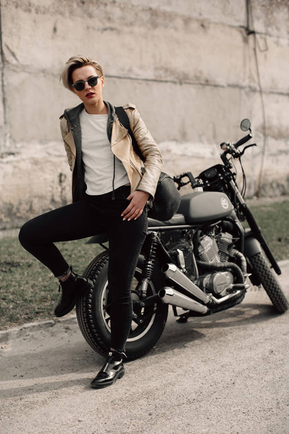 Une femme assise sur sa moto. Moto près d'un mur gris. fille aux cheveux courts. fille en lunettes noires et une veste en cuir. Vélo
 - Photo, image