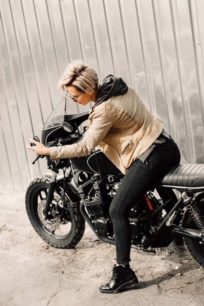 Woman sitting on her motorcycle. Motorcycle near a grey wall. girl looks at herself in the mirror of the motorcycle. girl with short hair. girl in black glasses and a leather jacket. Bike - Photo, Image
