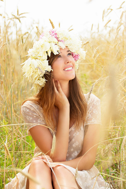 Alegria da natureza. Retrato de sorrindo bela jovem vestida de vestido branco e com flores na cabeça está sentado no campo de trigo amarelo
. - Foto, Imagem