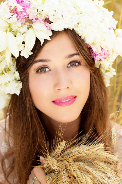 Pure beauty. Closeup portrait of beautiful young woman with flowers on her head keeping wheat in her hands while posing in the yellow wheat field. - Foto, imagen