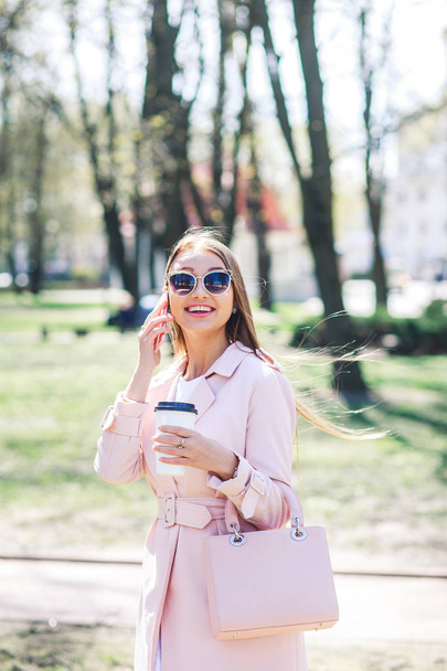 Fashionable woman with phone and cofee in the city. Fashion woman in a sunglasses and pink jacket outdoor - Foto, Bild