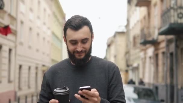 Happy beard young man texting on smartphone and drinking coffee in the street at sunset. He walks down the street, motion camera - Filmati, video