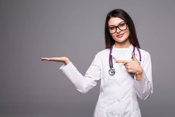 Female medical doctor showing copy space. Woman in white professional uniform with stethoscope on neck invite people to clinic hospital on grey. - Photo, Image