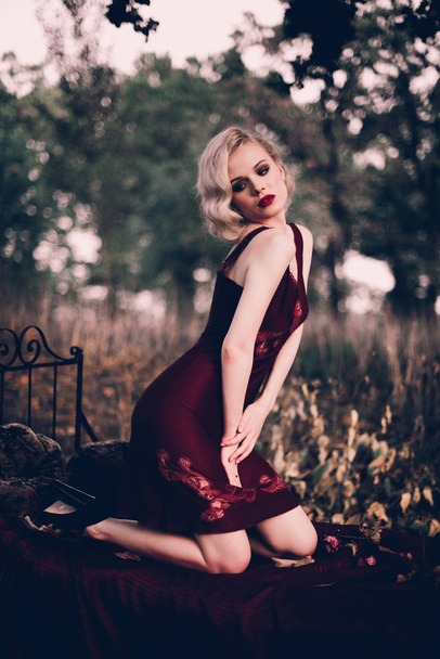 Όμορφο και κομψό ξανθιά γυναίκα με κόκκινα χείλη και τα μαλλιά τα κύματα φορώντας κρασί κόκκινο Νυχτικό ποζάρει στο κρεβάτι σε εξωτερικούς χώρους το φθινόπωρο, ρετρό vintage στυλ και τη μόδα. - Φωτογραφία, εικόνα