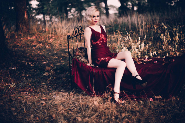 Belle et élégante femme blonde aux lèvres rouges et aux vagues de cheveux portant une nuisette rouge vin posant sur le lit en plein air en automne, style vintage rétro et mode
. - Photo, image