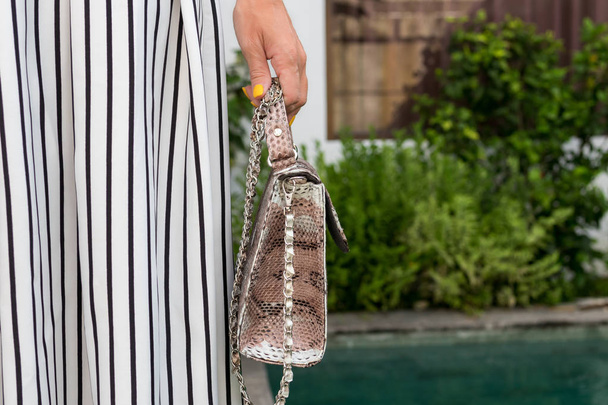 Fashionable woman holding luxury snakeskin python bag. Elegant outfit. Close up of purse in hands of stylish lady. Model posing near the swimming pool. Bali island. - Photo, Image