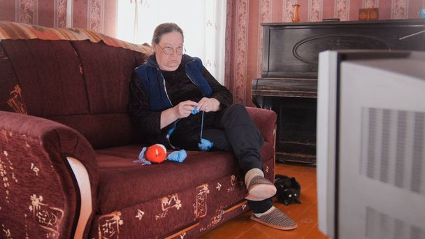 Oude dame thuis op de bank kijken naar Tv en breit wollen sokken - Foto, afbeelding