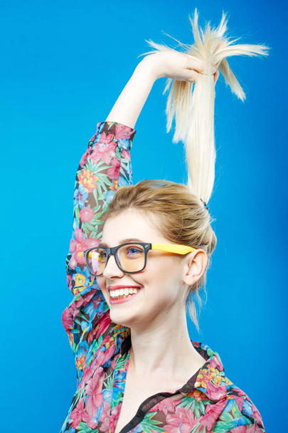 Веселая симпатичная девочка в модных очках - это Пепе в студии. Портрет смешной блондинки с хвостиком в разноцветной рубашке на синем фоне
. - Фото, изображение