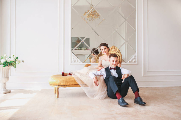 Bride in beautiful dress and groom in black suit sitting on sofa indoors in white studio interior like at home. Trendy wedding style . - Zdjęcie, obraz