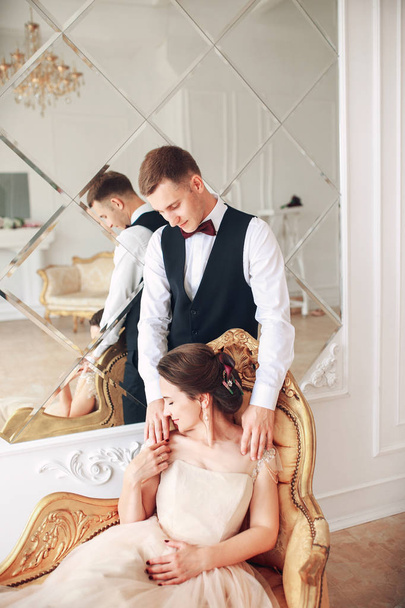 Bride in beautiful dress and groom in black suit sitting on sofa indoors in white studio interior like at home. Trendy wedding style . - Photo, image