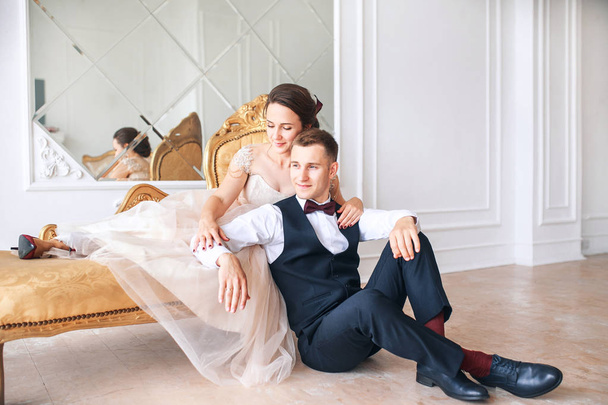 Bride in beautiful dress and groom in black suit sitting on sofa indoors in white studio interior like at home. Trendy wedding style . - Фото, зображення