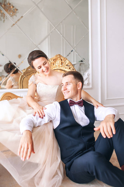 Bride in beautiful dress and groom in black suit sitting on sofa indoors in white studio interior like at home. Trendy wedding style . - Фото, изображение