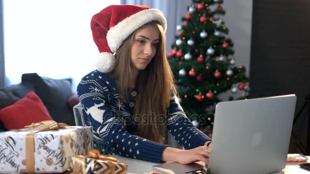Smiling young woman in santa claus chating with friend on laptop and drinking coffee on the christmas tree background in the living room. - Materiał filmowy, wideo