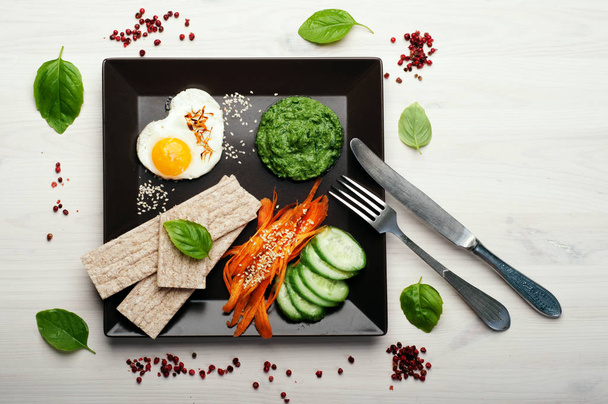 The concept of a delicious and healthy breakfast of a person who cares about his health. Fried egg, spinach puree, diet loaves, pieces of cucumber and carrots in a square plate on wooden background. - Foto, Imagem