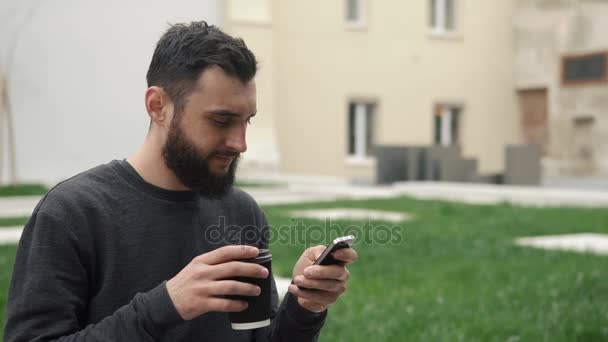 Happy beard young man texting on smartphone and drinking coffee in the street at sunset - Metraje, vídeo