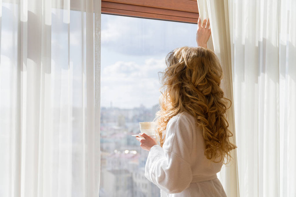 Beauty girl drinking coffee. Beautiful woman opening curtains, looking out the window and enjoying her morning coffee - Photo, image