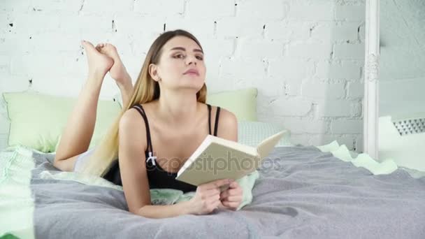 Young brunette woman reading a book on her bed and smiling - Imágenes, Vídeo