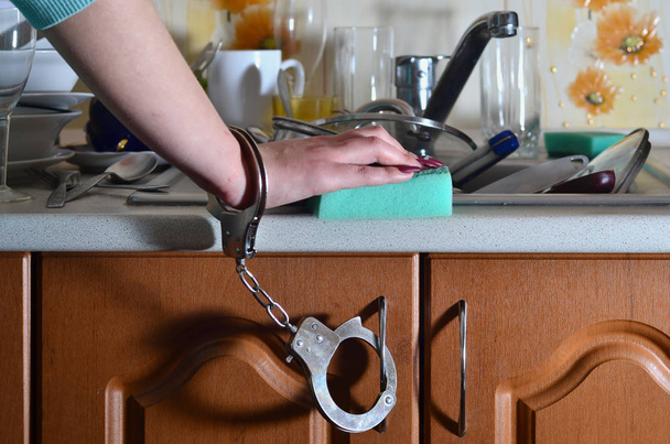 The hand of a young girl with a kitchen sponge, handcuffed to a kitchen counter with a lot of unwashed dishes, plates, cups and cutlery. The concept of forcing women to work in the kitchen - Photo, Image