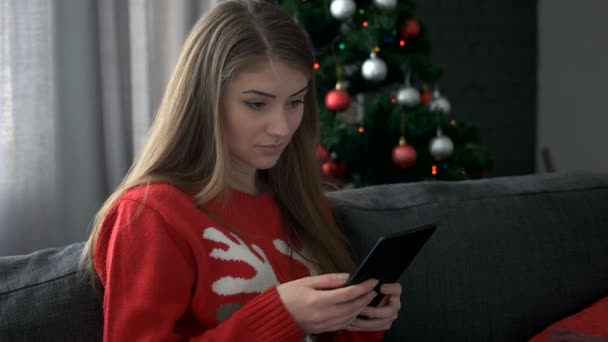 Portrait of a young attractive woman wearing sweater with deer sitting on sofa and using tablet on Christmas tree background. - Video