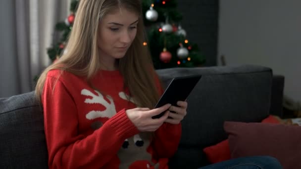 Young attractive woman wearing sweater with deer sitting on sofa and chating with friends in tablet on Christmas tree background. - Video