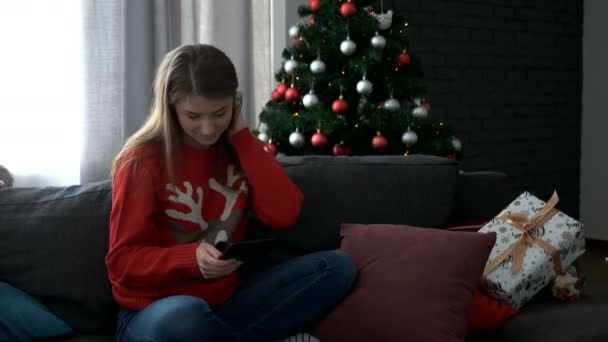 Jonge aantrekkelijke vrouw komt naar de woonkamer, zittend op de Bank met gave en start met het gebruik van Tablet PC op kerstboom achtergrond. - Video