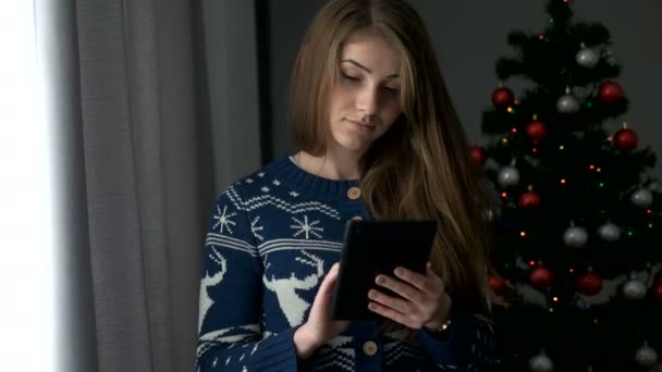 Portret van jonge aantrekkelijke vrouw die op tablet op de achtergrond van het venster met de kerstboom te typen. Close-up. - Video