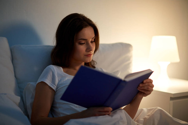 young woman reading book in bed at night home - 写真・画像