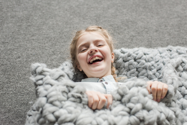 adorable girl laughing and lying on floor with grey knitted blanket - Фото, изображение