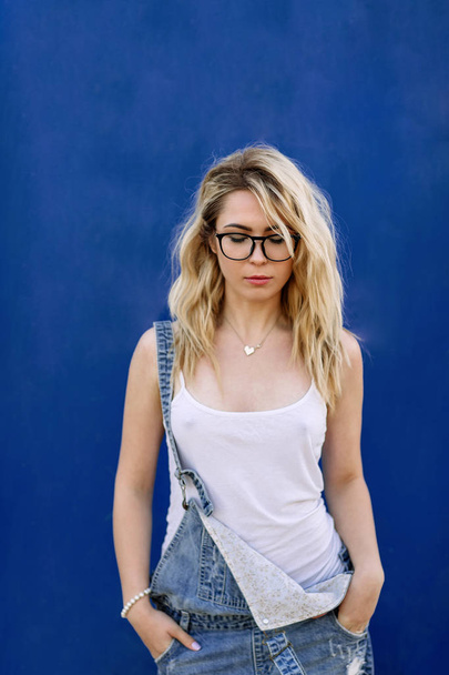 Young stylish blond girl in denim overalls made of denim and on a blue background - Foto, Bild