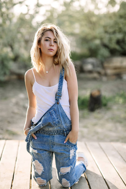 Young stylish blonde girl in denim overalls outdoors with natural daylight, a sunny day - Foto, Imagem