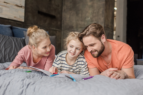 Happy parents lying on bed and looking at smiling daughter drawing - Фото, зображення