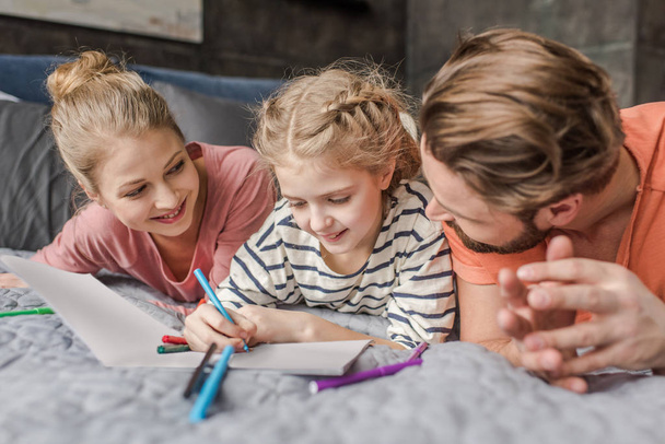 Happy parents lying on bed and looking at smiling daughter drawing - Foto, Bild